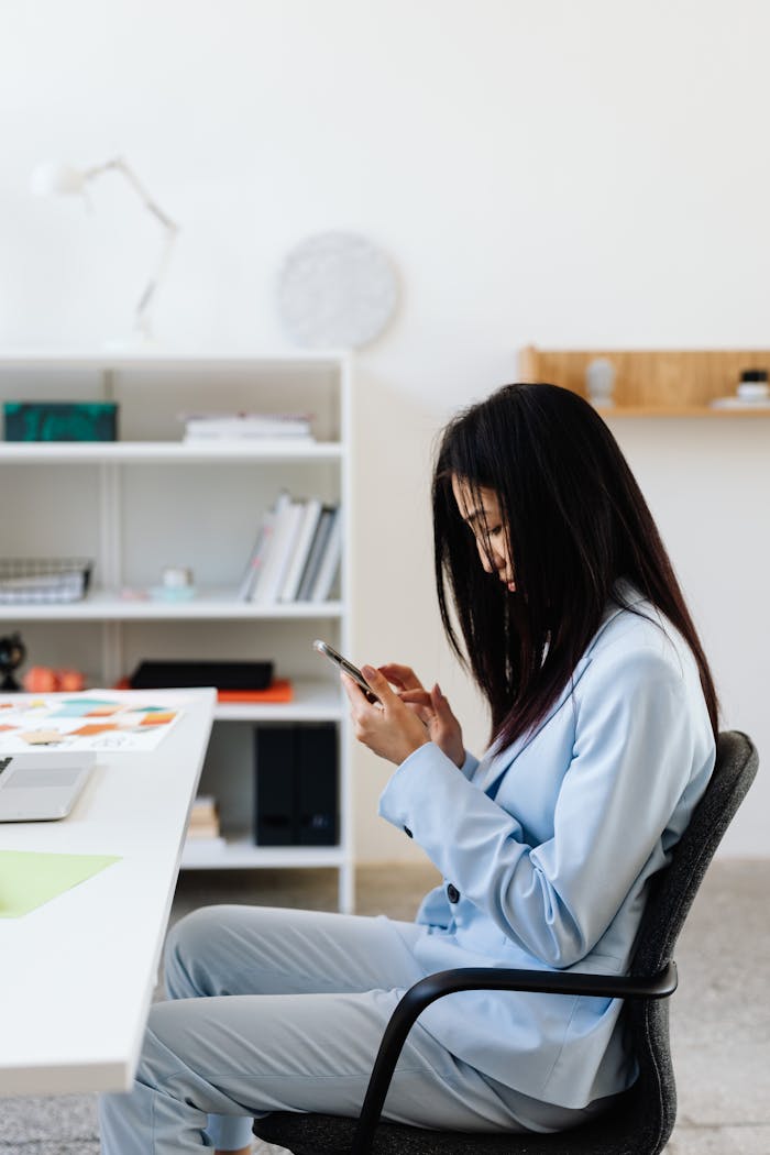 Businesswoman in a blue suit using smartphone while seated in a modern office workspace.